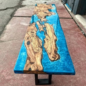 May include: A wooden table top with a blue epoxy resin inlay. The resin has a swirling pattern that resembles water. The table is supported by a black metal base.