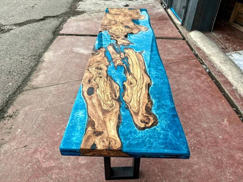 May include: A wooden table top with a blue epoxy resin inlay. The resin has a swirling pattern that resembles water. The table is supported by a black metal base.