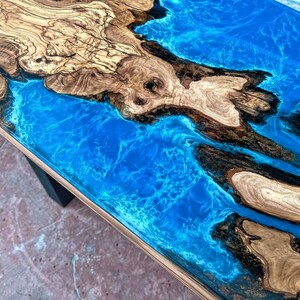 May include: A close-up of a wooden table top with a blue epoxy resin river running through it. The wood has a natural grain pattern and the resin has a swirling effect.