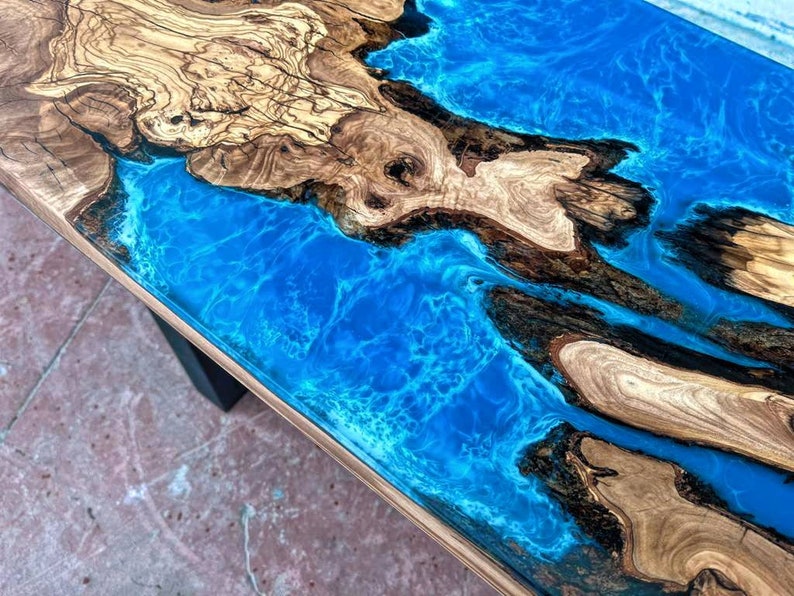 May include: A close-up of a wooden table top with a blue epoxy resin river running through it. The wood has a natural grain pattern and the resin has a swirling effect.