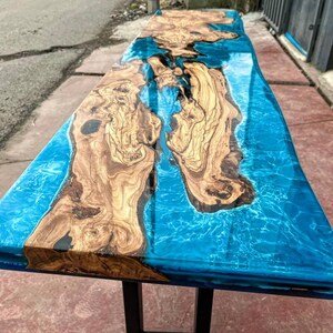 May include: A long, wooden table top with a blue epoxy resin inlay. The resin has a swirling pattern that resembles water. The table top is supported by black metal legs.
