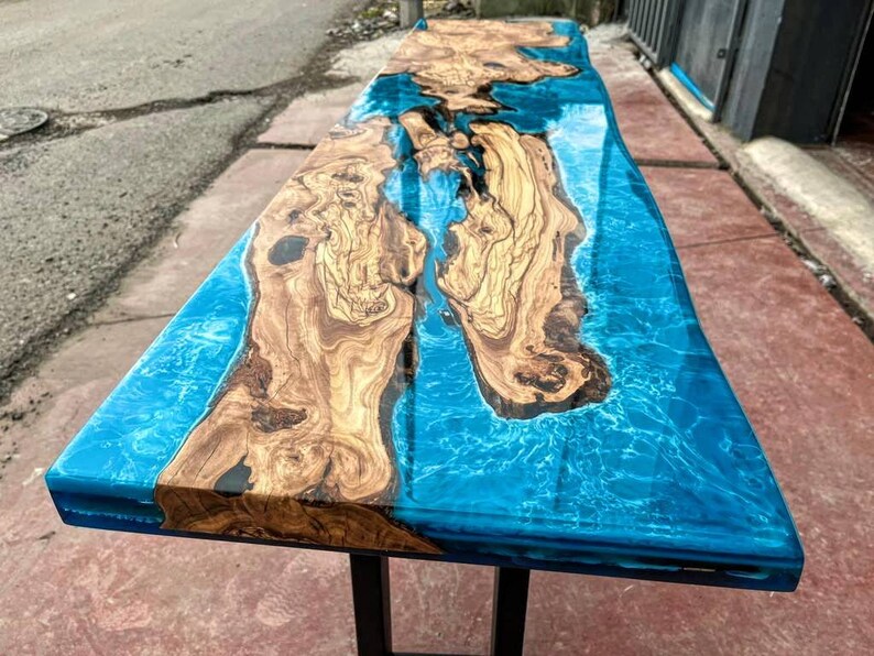May include: A long, wooden table top with a blue epoxy resin inlay. The resin has a swirling pattern that resembles water. The table top is supported by black metal legs.