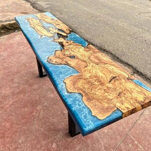 May include: A long, wooden table top with a blue epoxy resin inlay. The resin has a swirling, marbled effect. The table top is supported by a black metal frame.