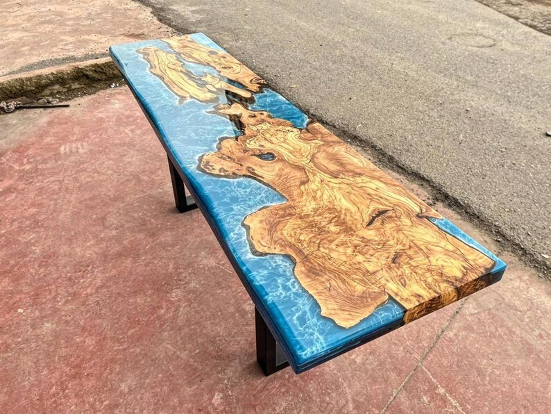 May include: A long, wooden table top with a blue epoxy resin inlay. The resin has a swirling, marbled effect. The table top is supported by a black metal frame.
