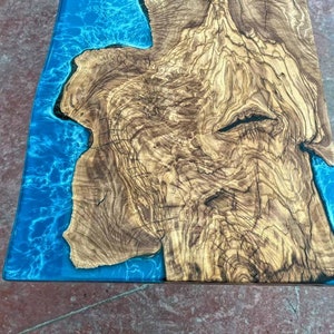 May include: A close-up of a wooden table top with a blue epoxy resin inlay. The wood has a natural, rustic look with lots of knots and grain. The epoxy resin is clear and has a slight blue tint. The table top is perfect for a modern or rustic home.