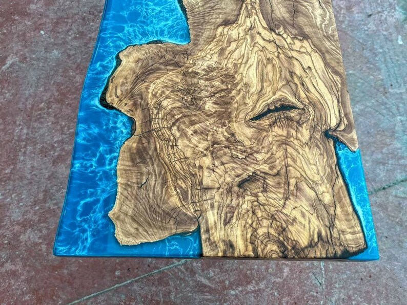 May include: A close-up of a wooden table top with a blue epoxy resin inlay. The wood has a natural, rustic look with lots of knots and grain. The epoxy resin is clear and has a slight blue tint. The table top is perfect for a modern or rustic home.