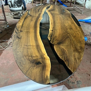 Könnte beinhalten: Ovaler Holztischplatte mit einem schwarzen Epoxidharzfluss, der durch die Mitte verläuft. Das Holz hat eine satte braune Farbe und ein natürliches Maserungsmuster.