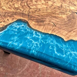 May include: Close-up of a wooden table top with a blue and white epoxy resin inlay. The resin has a swirling pattern that resembles water.