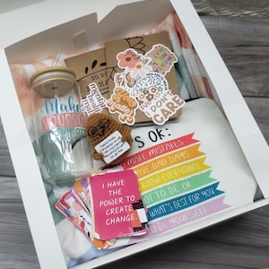 May include: A gift box filled with self-care items. Includes a glass tumbler with "Make Yourself a Priority" text, stickers, a crocheted owl, cards with positive affirmations, and a white t-shirt with a rainbow design and text.