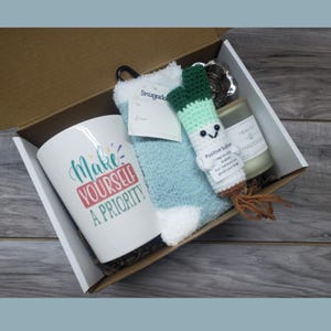 May include: A gift box containing a white mug with the text "Make Yourself A Priority", a pair of blue fuzzy socks, a crocheted Positive Scallion, a candle, and a tag. The box is open, revealing the contents.