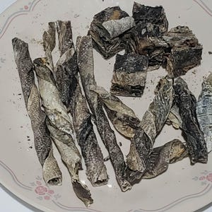 May include: A plate of dried fish snacks. The snacks are twisted and cut into various shapes, with a mix of gray, black, and tan colors. The plate has a floral design around the rim.