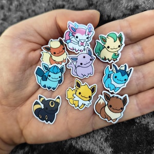 Puede incluir: Una colección de pegatinas de personajes de dibujos animados coloridos con varios diseños. Las pegatinas muestran una variedad de criaturas en tonos de azul, amarillo, marrón, rosa, verde y morado. Cada pegatina tiene un borde blanco.