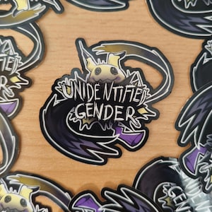 Könnte beinhalten: Ein Aufkleber mit dem Text "UNIDENTIFIED GENDER" in einer stilisierten Schriftart, der eine Zeichentrickfigur mit gelbem Kopf und schwarzen Akzenten zeigt. Der Aufkleber hat ein schwarzes und violettes Design.