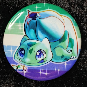 Może przedstawiać: Okrągła przypinka z postacią z kreskówki Bulbasaur w odcieniach zieleni i niebieskiego. Tło ma gradient kolorów, w tym zielony, biały, fioletowy i niebieski, z błyszczącymi akcentami gwiazd. Przypinka ma błyszczące wykończenie. Średnica ok. 5 cm.