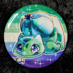 Peut inclure: Un bouton rond représentant un personnage de dessin animé Bulbasaur en vert et bleu, sur un fond aux couleurs de l'arc-en-ciel. Le bouton a une finition brillante avec des accents d'étoiles holographiques.