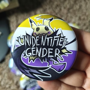 Könnte beinhalten: Ein runder Button mit einem Design der Non-Binary-Pride-Flagge. Der Button zeigt eine Cartoon-Figur und die Worte "UNIDENTIFIED GENDER" in einer schwarzen Sprechblase. Der Hintergrund ist gelb, weiß, lila und schwarz.