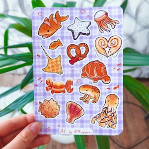 Puede incluir: Una hoja de pegatinas con ilustraciones de criaturas marinas y alimentos, como un pez payaso, una estrella de mar, una medusa y un pretzel. Los diseños son en naranja, blanco y marrón, sobre un fondo a cuadros morado y blanco. Firma del artista en la parte inferior.
