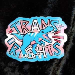 May include: A sticker with a blue dinosaur and the words "TRANS RIGHTS". The design uses pink and blue colours, with a white border and star accents. The sticker is on a black background.