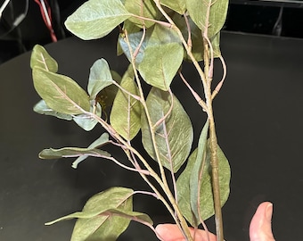Op de afbeelding: Een decoratieve kunstmatige eucalyptus-tak met meerdere gedempte groene bladeren met zichtbare roze-bruine nerven en stelen. De tak bevat kleine trossen van piepkleine, lichtgekleurde knoppen aan de uiteinden, wat een realistische botanische structuur laat zien.