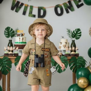 May include: A young child in a khaki safari outfit, complete with a hat and binoculars, stands in front of a "Wild One" banner. The background features balloons in green, gold, and leopard print, and a cake with animal decorations.