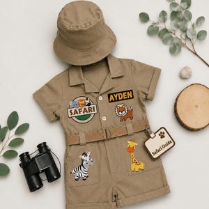 May include: A tan short-sleeved romper with a matching bucket hat, featuring embroidered patches of a lion, zebra, and giraffe, the text "SAFARI" and "AYDEN", and a "Safari Guide" badge, paired with black binoculars.