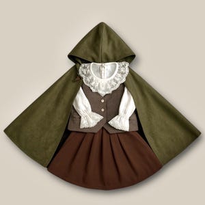 May include: A child's costume comprising a dark olive green hooded cape, a brown buttoned waistcoat, a white blouse with lace detailing, and a brown pleated skirt. The outfit is styled to resemble historical clothing.