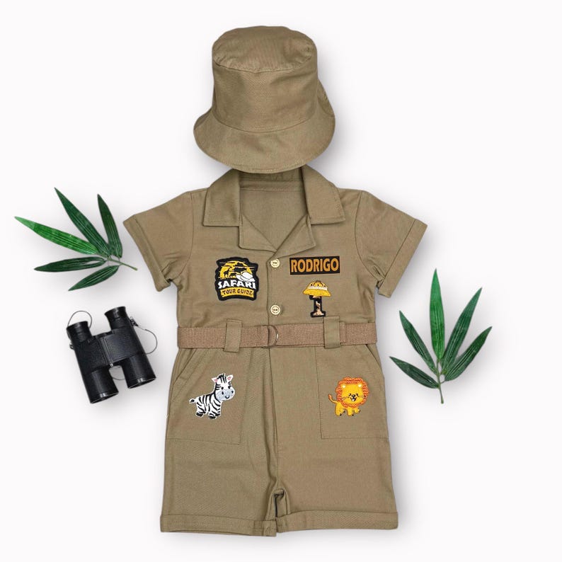 May include: A khaki safari-themed outfit for children, including a short-sleeved jumpsuit with a belt, a matching bucket hat, and a pair of black binoculars. The jumpsuit has patches with animal designs and the name "RODRIGO".