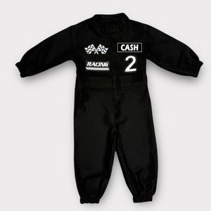 Op de afbeelding: Een zwarte jumpsuit met lange mouwen en witte patches op de borst. De patches bevatten twee gekruiste geblokte vlaggen, het woord RACING, de naam CASH en het cijfer 2. Het kledingstuk heeft een hoge halslijn en elastische mouwboorden.