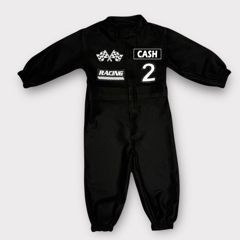 Op de afbeelding: Een zwarte jumpsuit met lange mouwen en witte patches op de borst. De patches bevatten twee gekruiste geblokte vlaggen, het woord RACING, de naam CASH en het cijfer 2. Het kledingstuk heeft een hoge halslijn en elastische mouwboorden.