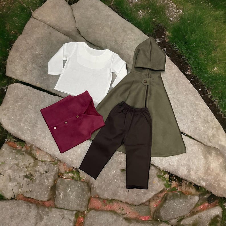 Puede incluir: Un conjunto de ropa que consta de una camisa blanca de manga larga, un chaleco burdeos oscuro con tres botones, un pantal&oacute;n marr&oacute;n oscuro y una capa con capucha de color verde oliva. Los art&iacute;culos est&aacute;n dispuestos sobre una superficie plana.