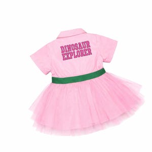May include: Pink collared dress with a green waist band and a layered tulle skirt. The back of the dress has the words "DINOSAUR EXPLORER" printed in pink.