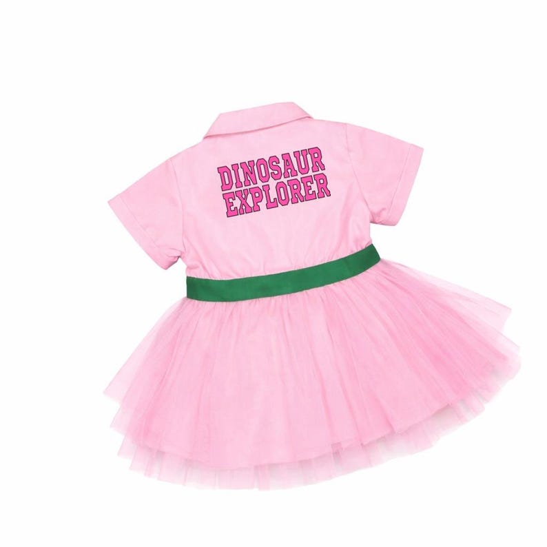May include: Pink collared dress with a green waist band and a layered tulle skirt. The back of the dress has the words "DINOSAUR EXPLORER" printed in pink.