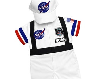 Może przedstawiać: Biały strój astronauty z motywem NASA dla dziecka, w tym czapka i kombinezon. Kombinezon ma czarne akcenty, imię "NOAH" i naszywkę z flagą USA. Czapka i kombinezon wyświetlają logo NASA.
