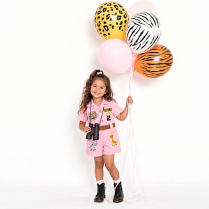 May include: A pink safari-themed romper featuring a brown belt, black binoculars, and patches including the name Stella, the number 2, a zebra, and a giraffe. The outfit is paired with black boots and a cluster of animal print and pink balloons.