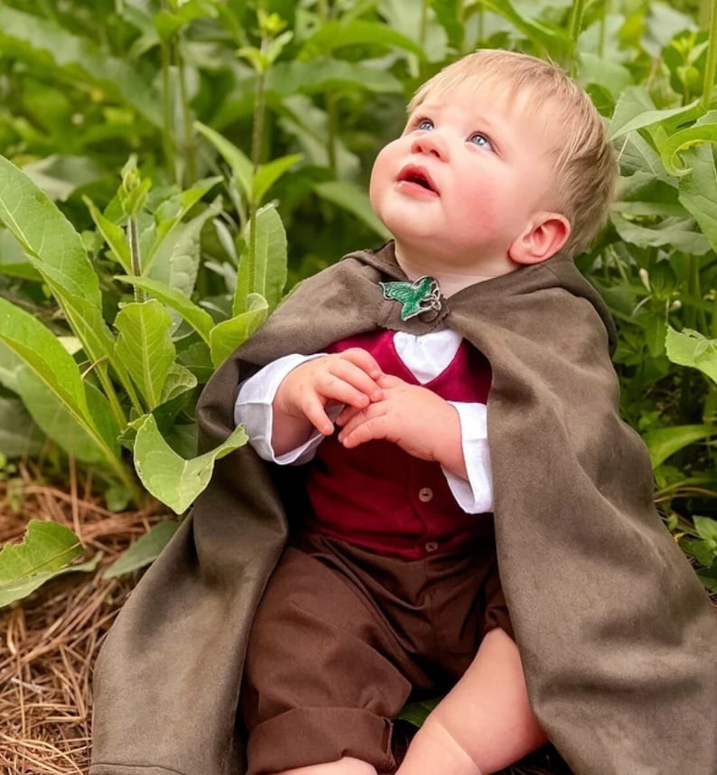 Puede incluir: Un ni&ntilde;o peque&ntilde;o con una capa con capucha de color marr&oacute;n oscuro sujeta con un broche en forma de hoja verde, una camisa blanca de manga larga, un chaleco abotonado de color rojo intenso y pantalones marrones. El conjunto tiene un dise&ntilde;o.