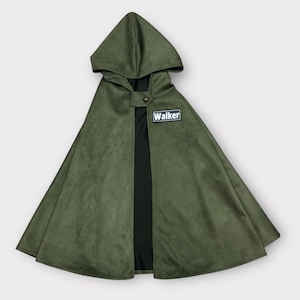 May include: An olive green hooded cape featuring a black interior lining and a single button closure at the neck. A rectangular black patch with a white border and the text "Walker" in white lettering is attached to the upper right chest area.