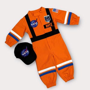 Può includere: Una tuta arancione a maniche lunghe con polsini elastici e cinghie verticali nere. Presenta un logo NASA, una toppa Space Explorer, una bandiera americana e un'etichetta nera con il nome RHYZ. È incluso un berretto nero con il logo della NASA.