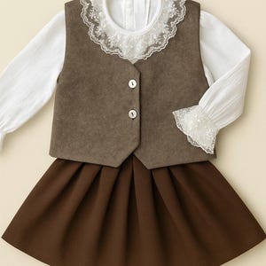 May include: A child's three-piece outfit comprising a white blouse with lace collar and cuffs, a brown waistcoat with two buttons, and a brown pleated skirt. The ensemble is designed for a young child.