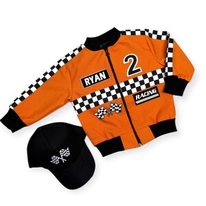 Può includere: Giacca da corsa arancione con dettagli a scacchi bianchi e neri, il numero 2 e il nome "RYAN". Include un berretto da baseball nero con grafica a bandiera a scacchi. La giacca ha una chiusura con cerniera e polsini neri.