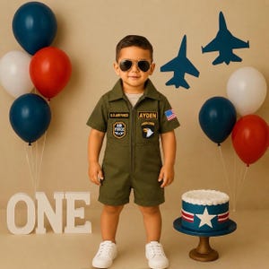 Peut inclure: Une combinaison courte vert olive à manches courtes avec fermeture éclair frontale, col et écussons brodés incluant U.S. Air Force, un emblème d'aigle et un petit drapeau américain sur la manche, avec le nom Ayden imprimé sur un écusson.