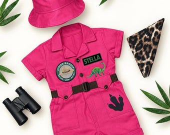Puede incluir: Un mameluco rosa brillante de manga corta con cinturón marrón, sombrero de pescador a juego y pañuelo con estampado de leopardo. El mameluco incluye un parche de Junior Ranger, un dinosaurio verde, una huella negra y el nombre STELLA.