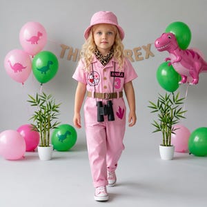 May include: A pink jumpsuit with short sleeves, a brown belt, and a leopard print neck scarf. The outfit features a circular patch with a dinosaur, a black name tag reading ANIKA, and the number 3. Black binoculars hang around the neck.