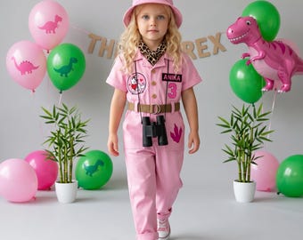 Puede incluir: Un mono rosa con sombrero de pescador a juego, que incluye un pañuelo con estampado de leopardo, un cinturón marrón y binoculares negros. El mono presenta un parche circular de dinosaurio, una etiqueta con el nombre ANIKA y un número 3 rosa.
