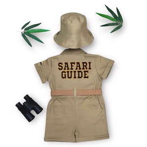 May include: A khaki-colored safari guide costume set, including a short-sleeved jumpsuit with "SAFARI GUIDE" printed on the back, a matching hat, and a belt. The set is accompanied by a pair of black binoculars. Green leaves are placed on the top and left side.