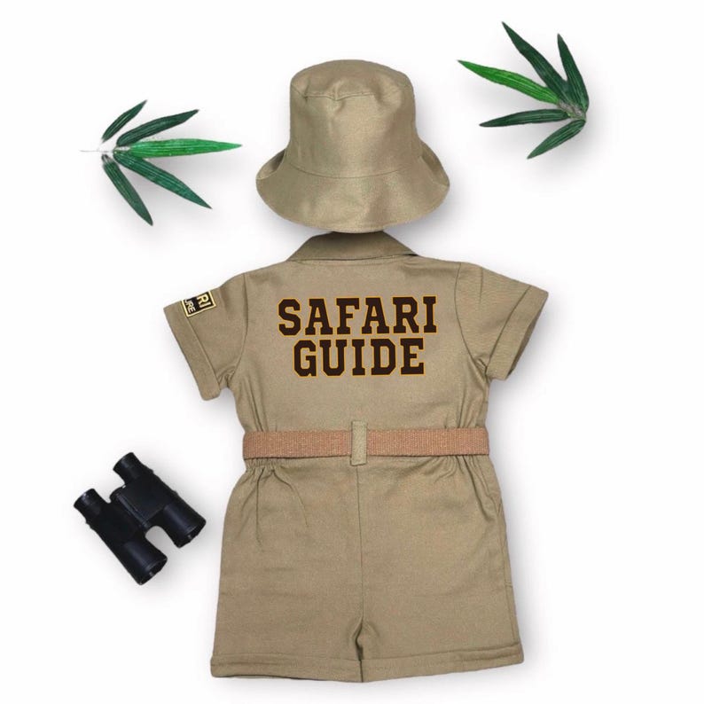 May include: A khaki-colored safari guide costume set, including a short-sleeved jumpsuit with "SAFARI GUIDE" printed on the back, a matching hat, and a belt. The set is accompanied by a pair of black binoculars. Green leaves are placed on the top and left side.