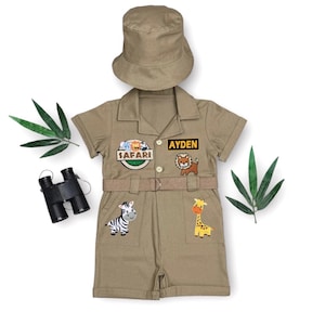 May include: Khaki safari-themed outfit for kids, including a short-sleeved shirt and shorts set, a matching hat, and a belt. The shirt features the name "Ayden" and safari patches with animal illustrations. A pair of binoculars is also included.