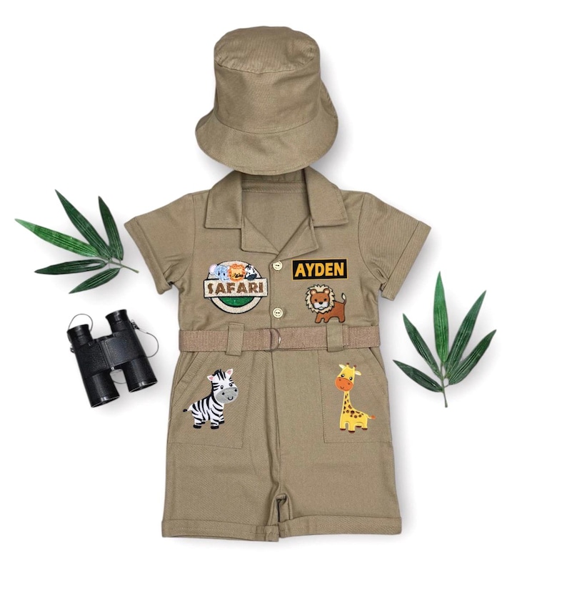 May include: Khaki safari-themed outfit for kids, including a short-sleeved shirt and shorts set, a matching hat, and a belt. The shirt features the name "Ayden" and safari patches with animal illustrations. A pair of binoculars is also included.