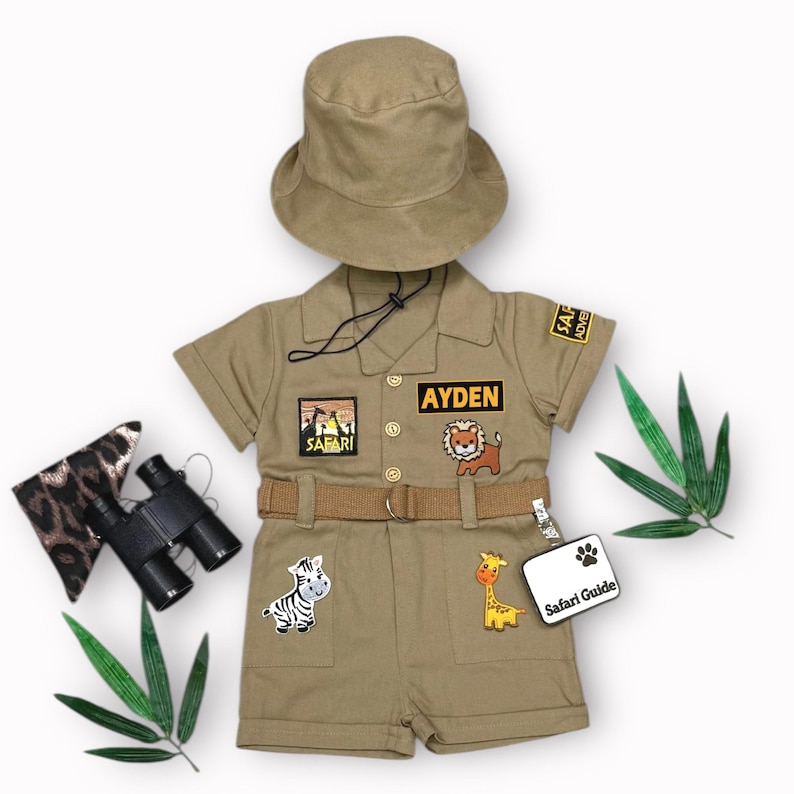 May include: Khaki safari outfit set with matching hat, belt, and patches. Includes a shirt and shorts with animal-themed patches, a name patch that says "AYDEN", and a "Safari Guide" tag. Accessories include binoculars and a leopard print pouch.