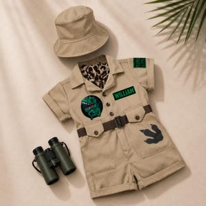 Può includere: Una tutina beige a maniche corte con abbottonatura frontale, cintura marrone e cappello da pescatore. La tutina presenta una toppa con dinosauro verde, una toppa nera con il testo WILLIAM, una toppa DINO EXPLORER e una stampa di impronta.