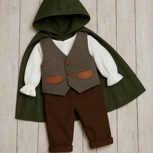 May include: A dark green hooded cape worn over a white long-sleeved shirt with ruffled cuffs, a brown vest featuring two brown buttons and two tan pocket flaps, and solid brown trousers with cuffed hems, arranged as a complete outfit set.